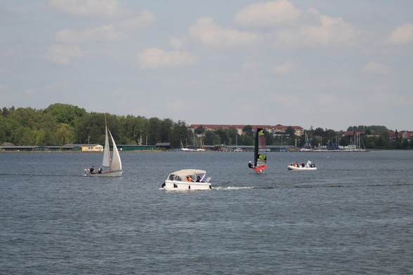 Auf einem großen Gewässer fahren Boote und Surfer mit und ohne Segel. Im Hintergrund kleine Hafenanlagen und Häuser.