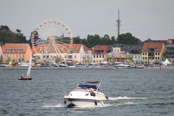 Im Vordergrund Boot und kleines Segelboot. Dahinter Hafenbereich mit Promenade, auf der Riesenrad und Karussel stehen. Dahinter Häuser, Bäume und ein Turm.