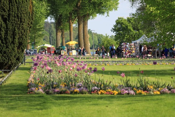 Blumenbeete mit blühenden Pflanzen im Vordergrund mit Rasen und Koniferen links. Dahinter Verkaufsstände und Leute auf den Weg.