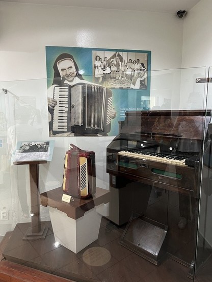 Museum display/shrine behind plexiglass, showing images of Irma Dulce playing her accordion. Below the picture is a real piano, and a small table holding a red piano accordion!