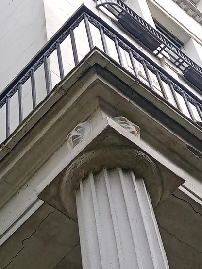 Blick auf ein weiß graues Haus, dessen Balkon von einer massiven Säule getragen wird und am Übergang von Säule zu Balkonbrüstung mit zwei maskenähnlichen Gesichtern geschmückt ist. Der Bildausschnitt ist so gewählt, dass die Brüstung des Balkons spitz auf den oberen Rand des Bildes zeigt. 