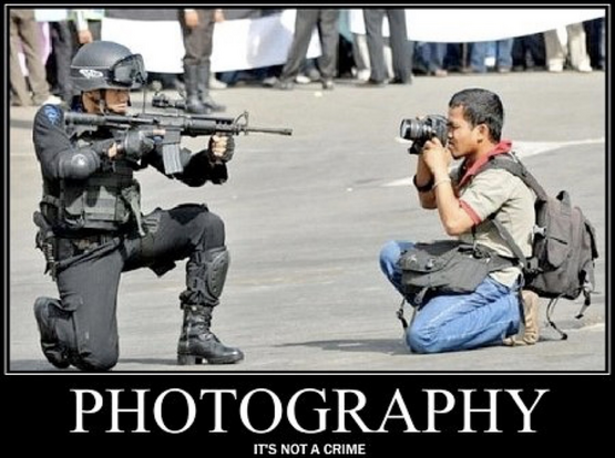 Image of a photographer kneeling and taking a photo of a soldier who is aiming a rifle. Below, the caption reads, "PHOTOGRAPHY: IT'S NOT A CRIME."