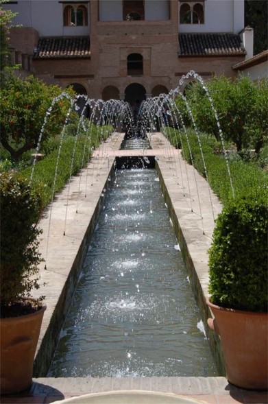 long pond, with stone sides, water arches fountains all the way to the end where there is a building with 3 arched doors and arched windows, Andalusia Style. Fruits trees on both sides of the long pond. 2 red stone pots with green buxus at the front on the left and right. 