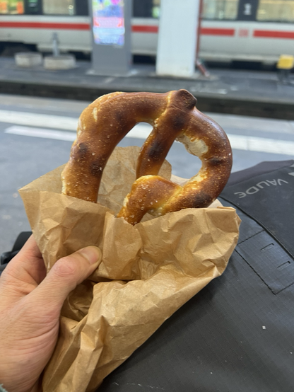 Gezeigt wird einen bretzel in einer Brötchentüte auf einem Rucksack, im Hintergrund ist ein Bahnhleis / Gleise zu sehen.  