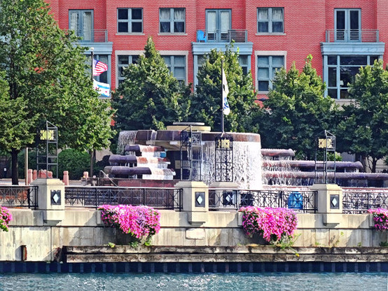 The Nicholas J Melas Centennial Fountain in Chicago IL, large multi-tiered fountain cascades water down its circular steps. Vibrant pink flowers hang from the railing enhancing the picturesque setting.