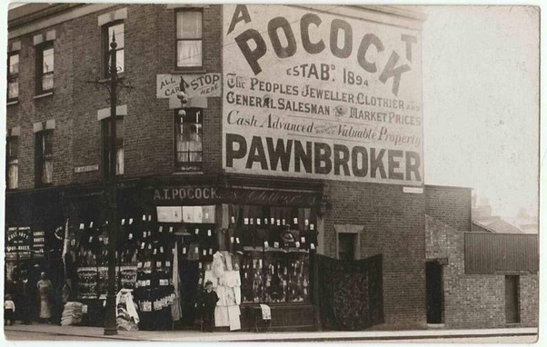 Black and white archival photo of a corner building with a shop on the ground floor with lots of goods on display and a person, presumably the shopkeeper, standing beside the door. 

There are various bits of signage on the building, but most prominent is a huge painted wall sign that fills an area the full width of the building by the upper two storeys. It has the name, 'A.T. Pocock' in huge letters at the top, and 'pawnbroker' in slightly smaller letters along the bottom. Between these are details of services and the date of establishment, 1894.
