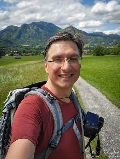 In this image, we see a middle-aged man taking a selfie outdoors in a grassy field with a cloudy sky in the background. The man is wearing sunglasses and a backpack, indicating he may be hiking or backpacking. He is smiling and posing for the selfie, showcasing a happy expression on his face. The dominant colors in the image are grey, with the accent color being a dark green. The man's face is the main focus of the image. The overall scene suggests a sense of adventure and travel, capturing a m…