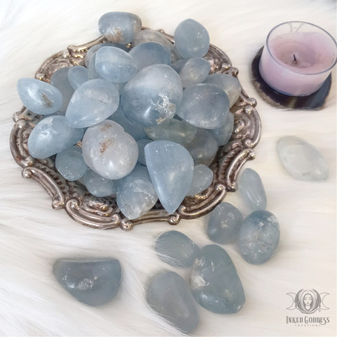 A number of "Celestite Tumbled Gemstone" specimens are arranged on a white faux-fur surface. A small silver bowl with ornate edges is filled with more gemstones. A tealight candle is shown for scale. Presented by Inked Goddess Creations.