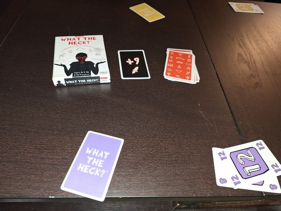 Photo of the card game What The Heck, aka Vulture Culture, during play.

The box depicting the silhouette of a male shrugging is visible next to the current point card, a positive 9, that is being bid on. Face down bid cards for the yellow and purple player are visible across the table from each other. 