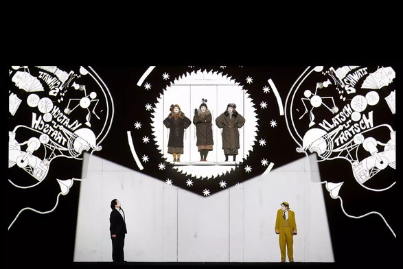 A still image from the performance of Mozart's The Magic Flute, as performed by San Francisco Opera. It shows three performers in the center in 1920's fashion, standing on platforms looking down two men in suits with faces painted white looking up. The entire background has two "phrenology-style head graphics showing the inner working of each brain with gears and words and details... out of the two eyes there is light projected down on the two actors standing on the stage floor. The entire backdrop is projected onto a flat black background which gives the stage a surreal multi-dimension. Quite a presentation to complement the fabulous music of the show.