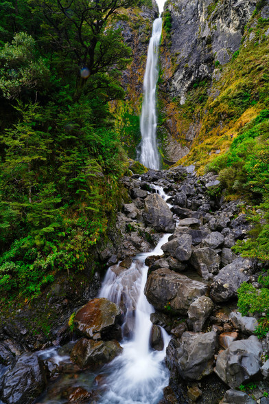 Devils Punchbowl Falls is one of the main tourist attractions in Arthur's Pass. At 130 metres high, with a clear view of the full length of the falls from the viewing platform, it's easy to see why. Water squirts out through an elevated narrow gorge at the top of the falls and drops into a small pool down below. The pouring of the "punch" is easy to see, but the circular pool or "punchbowl" is hidden behind a mound of boulders that block its view from the platform.
The walk to the falls is relatively short but quite steep and varied. It starts with a flat walk across the bridge over the Bealey River, followed by a narrower and higher suspension bridge across Devil's Punchbowl Creek. From there the walk heads uphill through a lush beech forest with amber mosses hanging suspended from the branches. There are numerous steps that will test your fitness before the track returns to gently undulating as you near the falls.