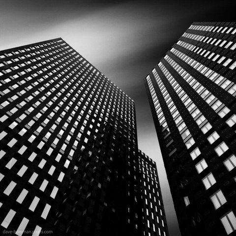 "Three Towers" is a black and white, long exposure photograph capturing three tall tower blocks in Amsterdam, Netherlands. The image looks up from the ground, highlighting the numerous windows that cover each building. The photograph has a semi-abstract quality, with the long exposure creating a smooth, almost surreal effect on the buildings' surfaces. The overall composition focuses on the stark geometry and towering presence of the structures against the sky.