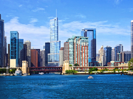 A vibrant Chicago cityscape features a variety of modern skyscrapers and high-rise buildings against a clear blue sky. The foreground is dominated by a calm body of water where boats can be seen.