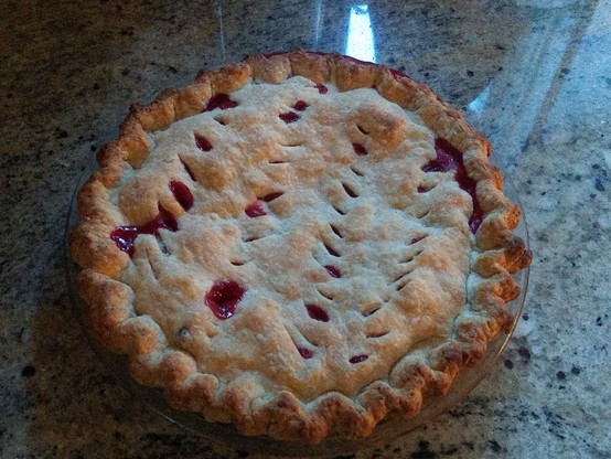 A pie cools in the counter. It's got a golden brown crimped crust around the edges and bits of red fruity filling have bubbled up.
