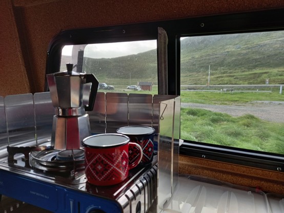 Zwei Blechtassen mit Norwegermuster stehen zusammen mit einem Espressokocher auf dem Gaskocher. Aus dem Fenster sieht man Grün und Berge. Das Bild entstand auf einem Campingplatz auf der Insel Magerøya in Nordnorwegen, auf der sich das Nordkap befindet.