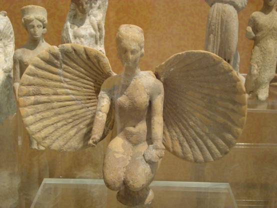 Terracotta figurine of Aphrodite kneeling in de nude, her left hand holding a libation bowl and her right hand missing. The clams of a sea shell spread behind her like wings.
