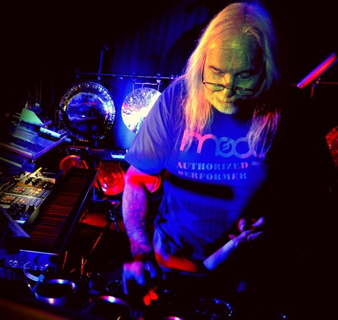 Bernd-Michael Land bei einem Liveauftritt. Er trägt langes graues Haar offen sowie eine Brille und ein T-Shirt mit dem Logo der Firma Moog. Im Vordergrund stehen Synthesizer. Im Hintergrund große Gongs.