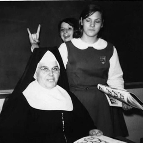 Black and white photo of two student girls in school uniforms standing behind the teacher (a nun wearing a habit), and one of the girls is making a a rock/devil horns sign behind the nun's head.