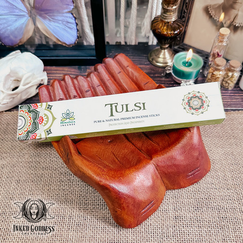 A rectangular box labeled "Tulsi Stick Incense" is laid across carved cupped wooden hands on a burlap-covered surface. Presented by Inked Goddess Creations.