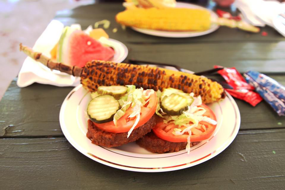 A picnic lunch plate with veggie burgers topped with tomatoes, lettuce, and pickles, accompanied by a delicious ear of fire-roasted corn on the cob.