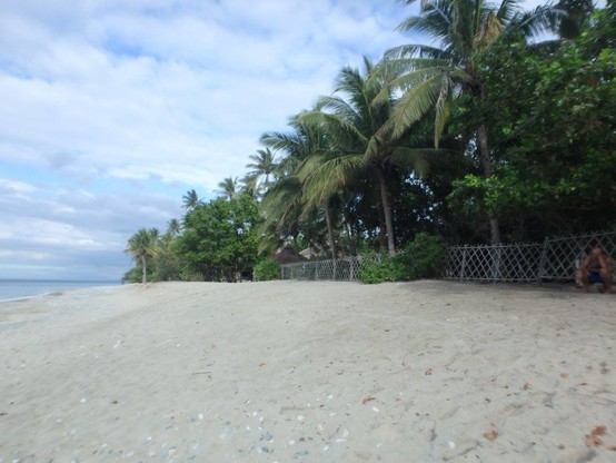 White beach, a glimpse of blue sea in the left of the photo to the center. The beach is fenced, the property next to the beach is private, Palmtrees on the property. the beach has white sand, some shells, and is deserted. The sky is blue with clouds smeared out over the blue like someone is icing a cake but has not finished yet. In the distance near the horizon the clouds get more grey. on the right in the corner is my friend, sitting at the fence. 