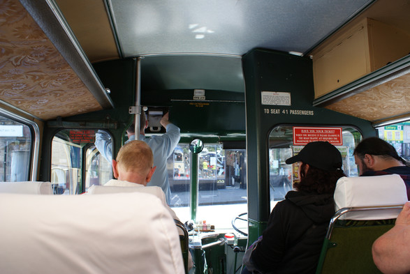 view from inside the bus about 4 rows in. Green liveried cloth back seats topped with white cotton. The driver is up front and is  changing the destination address.