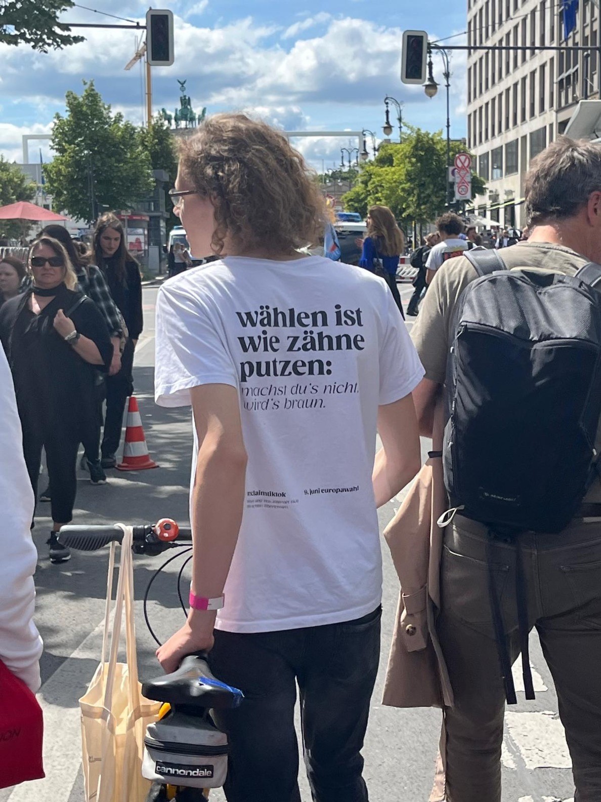 Person in a white T-shirt with German text, holding a bicycle and walking in a crowd on a city street. (Stemmen is als tandenpoetsen: doe je het niet dan wordt het bruin.)