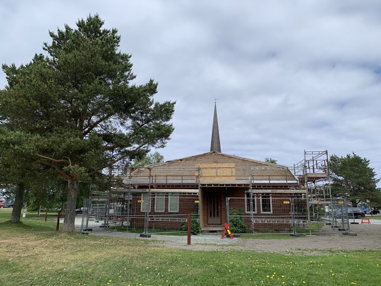 Den lokale kirka, Berg kirke, har fått fjernet all takstein. Det er stillas rundt kirka. Et høyt furutre til venstre for kirka.