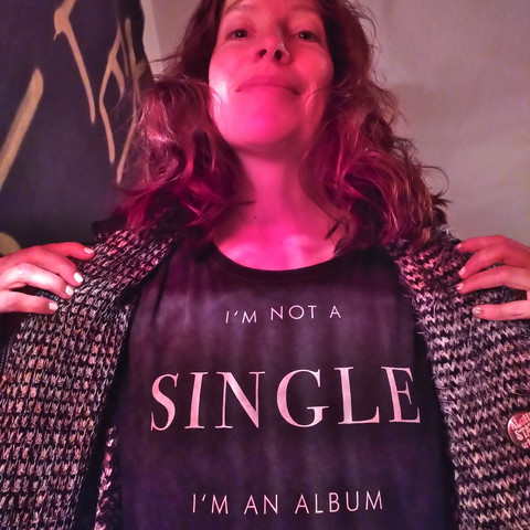 Fee in rotem Licht schräg von unten fotografiert. Auf ihrem T-Shirt steht: I'm not a single, I'm an album. Das Wort "single" ist um einiges größer gedruckt als der Rest der Schrift.