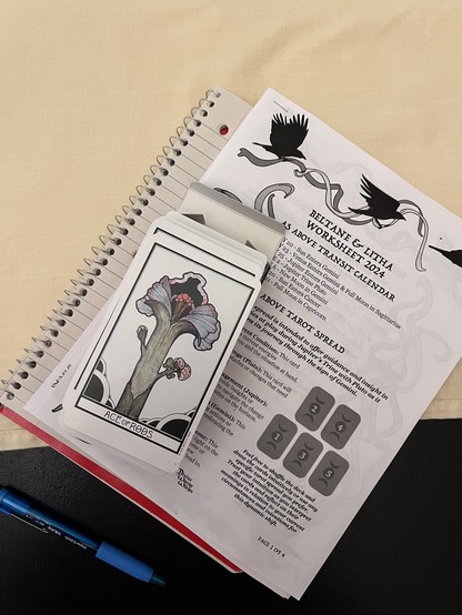 Photograph. A notebook, worksheet, and deck of tarot cards stacked on a cream-colored cloth on top of a black dining room table. A blue pen is visible at the bottom of the picture. The worksheet was provided as part of a celebration of Beltane & Litha and is decorated with images of crows and ribbons. It includes the As Above tarot spread mentioned in this post. The deck is David Palladini’s Aquarian Tarot. The only visible card is the Ace of Rods. 