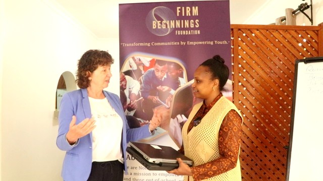 Flugpatin Anja L.-H. aus Breda (NL) brachte insgesamt 5 Laptops zur FIRM BEGINNINGS FOUNDATION in Tansania. Vielen Dank. Das Foto zeigt die Projektleiterin Joycelyne und Flugpatin Anja. Joycelyne hält 2 Laptops in ihren Händen. Beide stehen vor einem Roll-Up der tansanischen Projektes.
