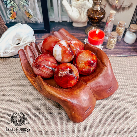 Six specimens of the "Carnelian Sphere" product are stacked inside a pair of carved cupped wooden hands. They are mostly deep red and orange with a bit of lighter material in some of them. Presented by Inked Goddess Creations.