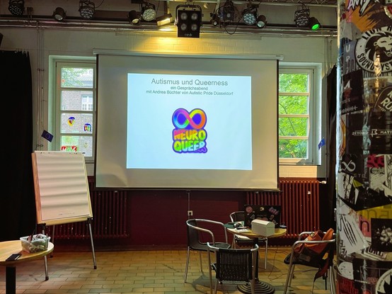 Raum vor der Präsentation zeigt einen Tisch mit Laptop und noch leeren Stühlen, sowie einen Screen mit Aufschrift "Autismus und Queerness" ein Gesprächsabend mit Andrea Büchter von Autistic Pride Düsseldorf" und dem regenbogenfarbenem Neuroqueer Zeichen.
