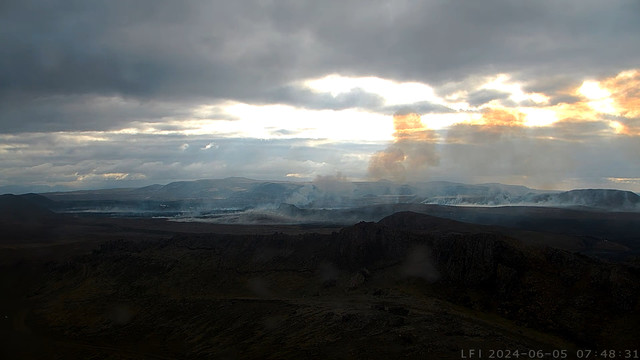 Foto, Weitwinkelaufnahme über das jüngste Eruptionsgebiet, starke Kontraste, dunkler Boden, in der Ferne graue Berge, dazwischen gelbgraue Rauchschwaden im Gegenlicht vor aufreißendem Himmel.