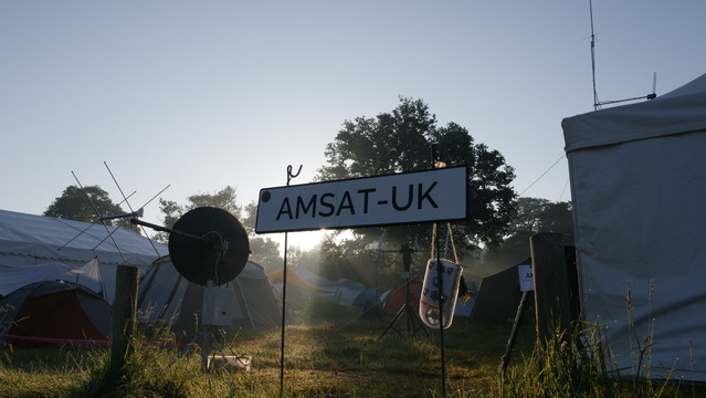 AMSAT-UK sign in the morning fog with the sun creating a nice streak of light behind it. Some antennas can be seen in view as well as out marquee tent