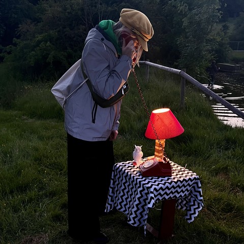 A person in a yellow hat holds the handset of an old rotary telephone to their ear in the dim light of dusk. The telephone sits on a small table with a tablecloth, a small white owl and a lit table lamp with a red shade. The table stands incongruously on the grass in front of a lake.