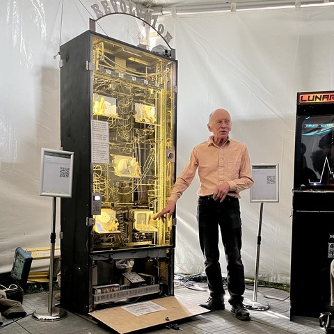 An incredibly famous man in shirt and jeans stands next to a towering mechanical contraption. Lit inside with yellow light, it is full of bent wire tracks for a giant ball bearing and five 3D tableaus He is pointing at the innards and talking about them.