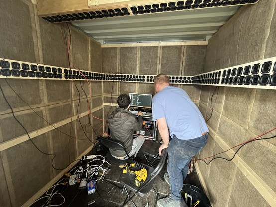 Two men are inside a small room baffled with sound dampening panels on the walls and surrounded by over 250 small speakers mounted on the sides and overhead. One of them sits at a computer while the other leans over his shoulder. There are wires and tools everywhere.