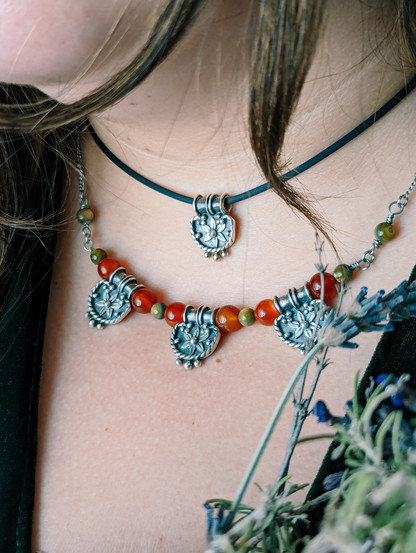 Dos collares: gargantilla con pequeño amuleto de inspiración íbera con una flor de 6 pétalos en el centro. Gargantilla de cuentas de ágata carneola y ágata verde con tres pequeños talismanes en forma de abanico con la misma flor central. En primer plano se adivina unas flores de lavanda y romero.