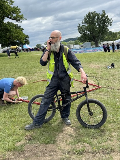 A bald man with an overly large grey beard, wearing manky blue overalls and a yellow hi-vis vest. He stands straddling a black BMX on the grass with a phone held against one ear. Behind can be seen tents and people.