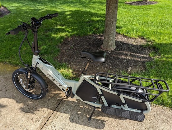 A picture of an Aventon Abound longtail cargo ebike on the edge of a sidewalk near some trees. The bike is sage green and features a step-through frame, storage bag, and smaller tires.