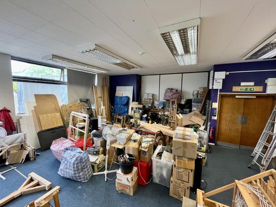 A large white room with an old blue carpet, photographed with a wide angle lens from one corner. There are piles of stuff in boxes everywhere. Bits of wood lean against the walls and a ladder is visible to one side.