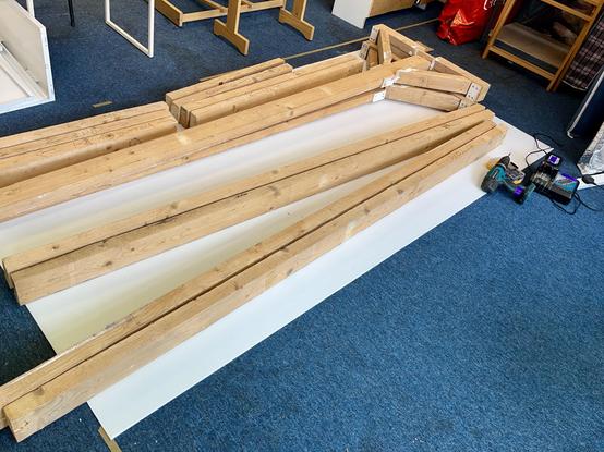 A pile of long lengths of timber lie on a white sheet of plastic on the blue floor. A power drill is off to one side.