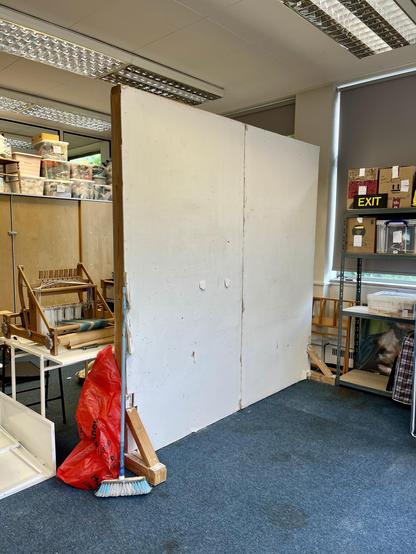 A blank white stud partition wall free-stands in the studio. A broom leans against it. Some shelves and cupboards can be seen around it with various bits on. A small weaving loom can be seen at the edge of the wall on a small table.