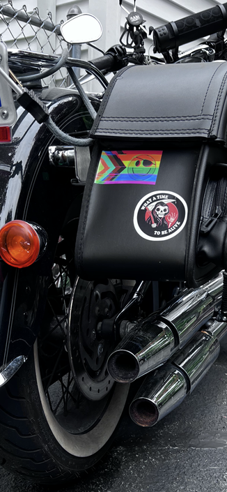 Picture of a saddlebag on a Harley, with a "Glitch Progress Pride" flag (A Progress Pride flag with "static" applied to it, a Happy face wearing an eyepatch) from essbee 
now keeping my "What A Time To Be Alive" sticker, from TheNib (RIP) featuring a Grim Reaper smiling (natch) and making an "OK" gesture company!