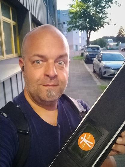 Joschi in a dark blue shirt on his way to the underground. He smiles at the camera with one eyebrow raised. He is holding a black roll-up banner with an orange and white Accessibility Club logo.