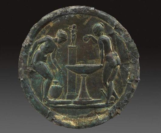 In the center is a large basin on a tapering pedestal. To the left, a nude woman with her hair wrapped in a cloth bends to pick up a water jar (kalpis) at the left. There are low bases or platforms below the kalpis and the basin. At the right, a nude woman with her hair pulled back tightly into a bun on the crown of her head leans on the basin with her left hand and pours oil into the basin from a bottle in her right. Behind her feet is an askos. Behind the basin is a pillar with mouldings abov…