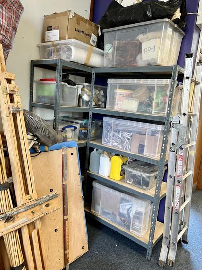 A corner of the studio with two sets of metal racked shelves. There are boxes piled up on all of the shelves. Bits of wood and an easel can be seen leaning in front of them.