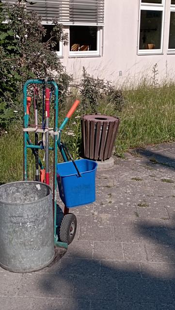 Sackkarre mit Zinkblech - Müllkübel, mit blauen Leergut-Sammeleimer, im Hintergrund ein Holzlatten-verkleideter Außen - Müllkübel, verschiedene Pflanzen, Schulgebäude 