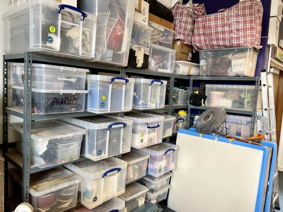 A fairly enormous pile of boxes and bags piled high on a corner entirely filled with shelves. Some large white and blue square things are leaning against one set. It all looks slightly dangerous, frankly.